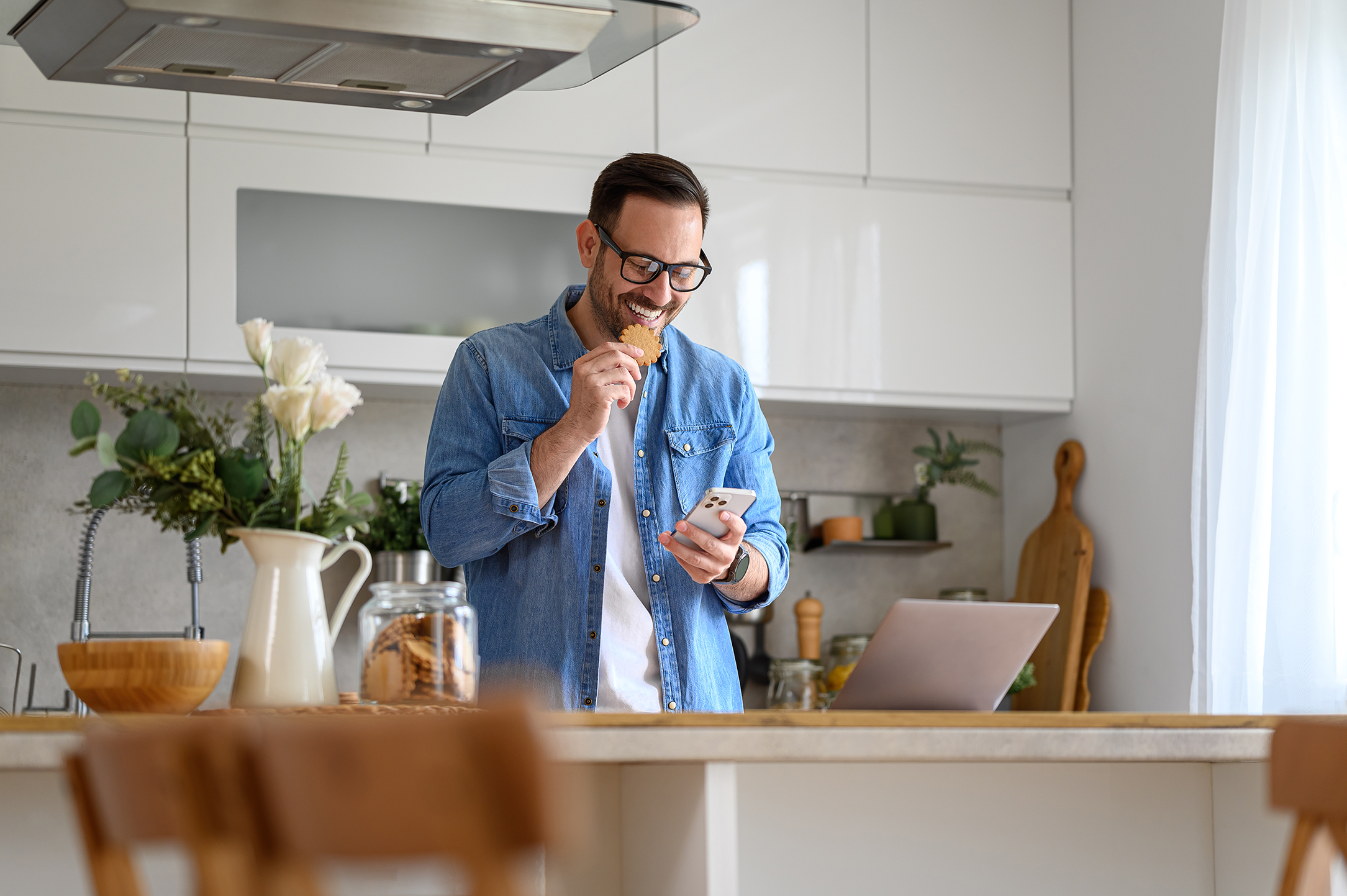 Man in kitchen eating a cookie while looking at his phone.